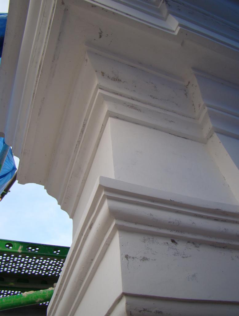 Detal architektoniczny kolumny z elementami sztukaterii, pomalowanej na biało, z widocznymi śladami patyny i rusztowaniem w tle.