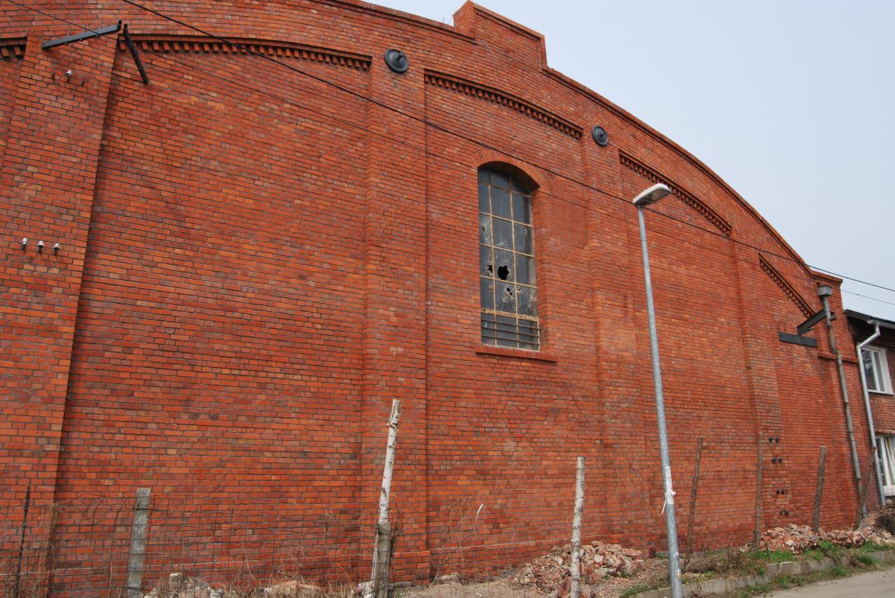 Ceglana ściana starego budynku z łukowym dachem i wybitymi szybami w oknie, widoczny fragment lampy ulicznej i ogrodzenia.
