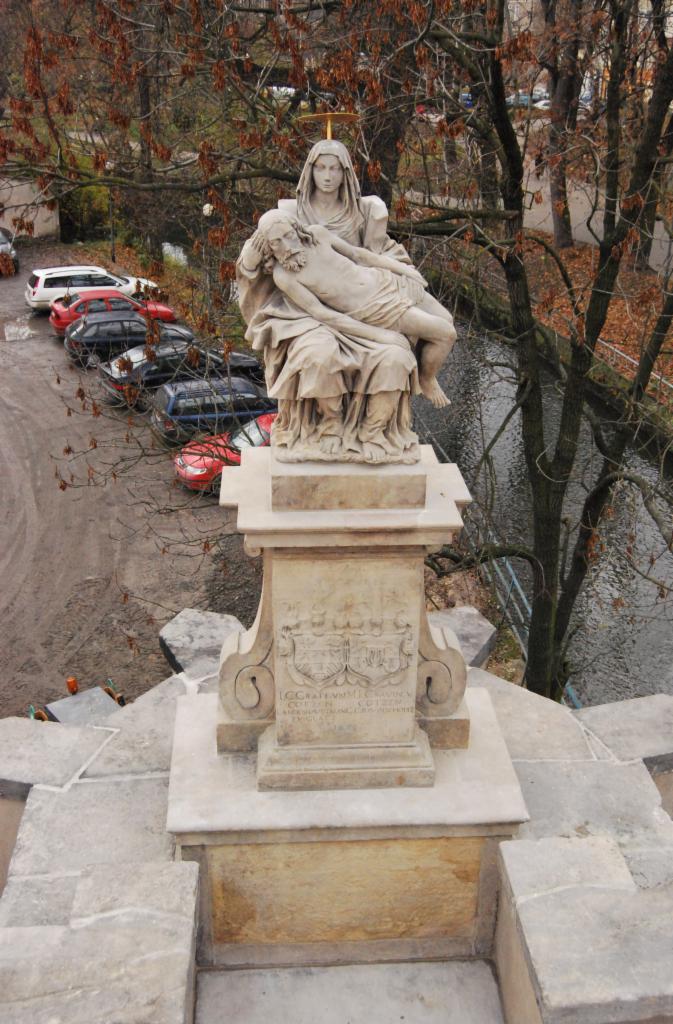 Kamienna rzeźba Piety na cokole z inskrypcją, widoczna z góry, z rzeką i parkiem w tle, oraz zaparkowanymi samochodami.