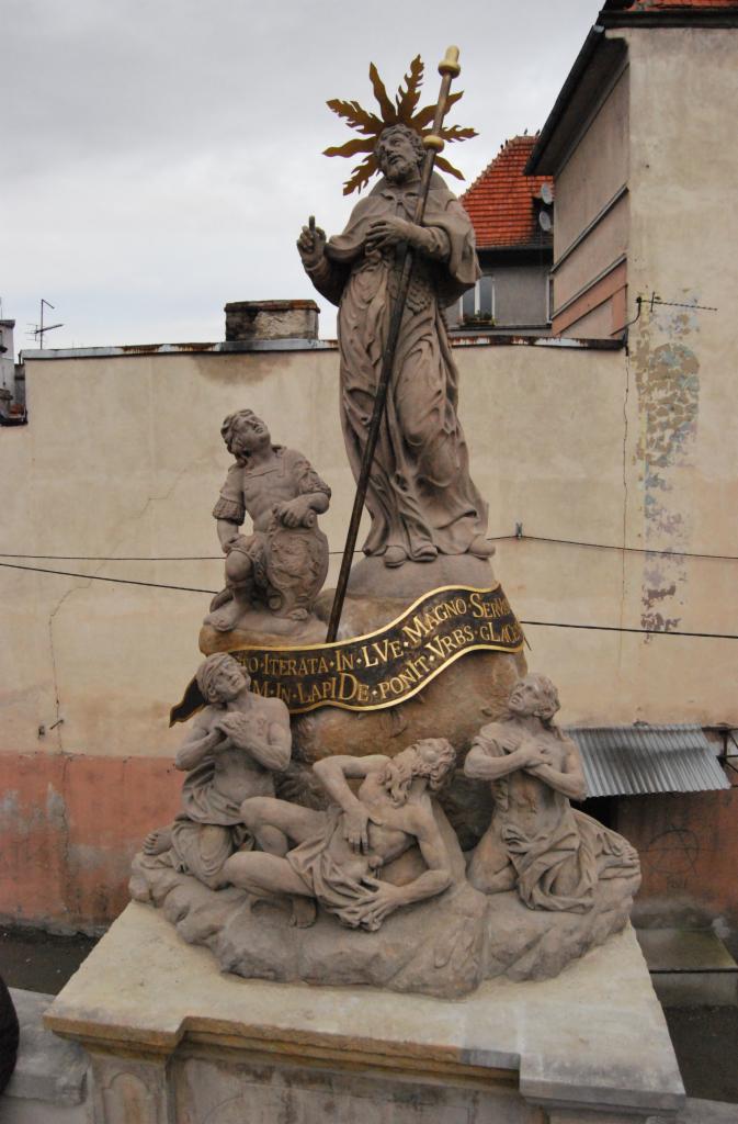 Kamienna rzeźba religijna na tle miejskiej zabudowy, przedstawiająca świętego z aureolą i laską, otoczonego przez klęczące postacie, z widoczną inskrypcją na wstędze.