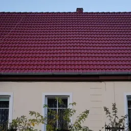 Czerwona dachówka na dwuspadowym dachu domu jednorodzinnego, widoczne okna, rynna, antena satelitarna i fragment elewacji.