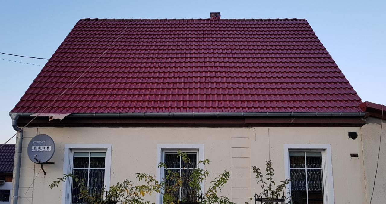 Czerwona dachówka na dwuspadowym dachu domu jednorodzinnego, widoczne okna, rynna, antena satelitarna i fragment elewacji.