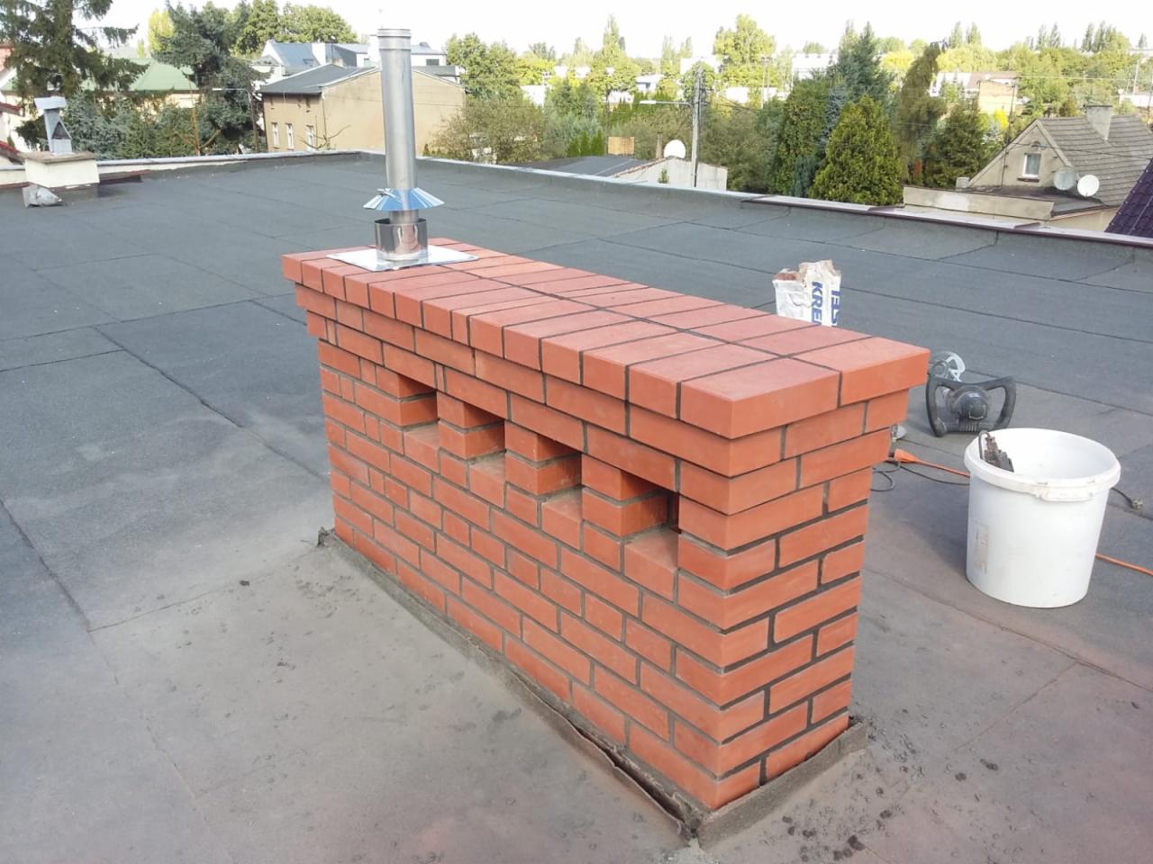 Ceglany komin z metalowym wkładem na dachu pokrytym papą, widoczne narzędzia i worek zaprawy murarskiej.