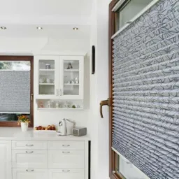 Plisowana osłona okienna z delikatnym, szarym wzorem kwiatowym w jasnej kuchni z białymi szafkami i drewnianą ramą okienną.