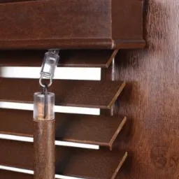 Szczegółowe ujęcie brązowych żaluzji drewnianych z widocznym mechanizmem sterującym na tle drewnianej ściany.