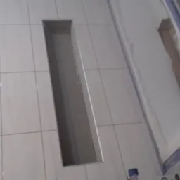 Ułożone jasne płytki podłogowe z wyciętym prostokątnym otworem, widoczny fragment brodzika z zabezpieczeniem folią i pojemnik z klejem w rogu.