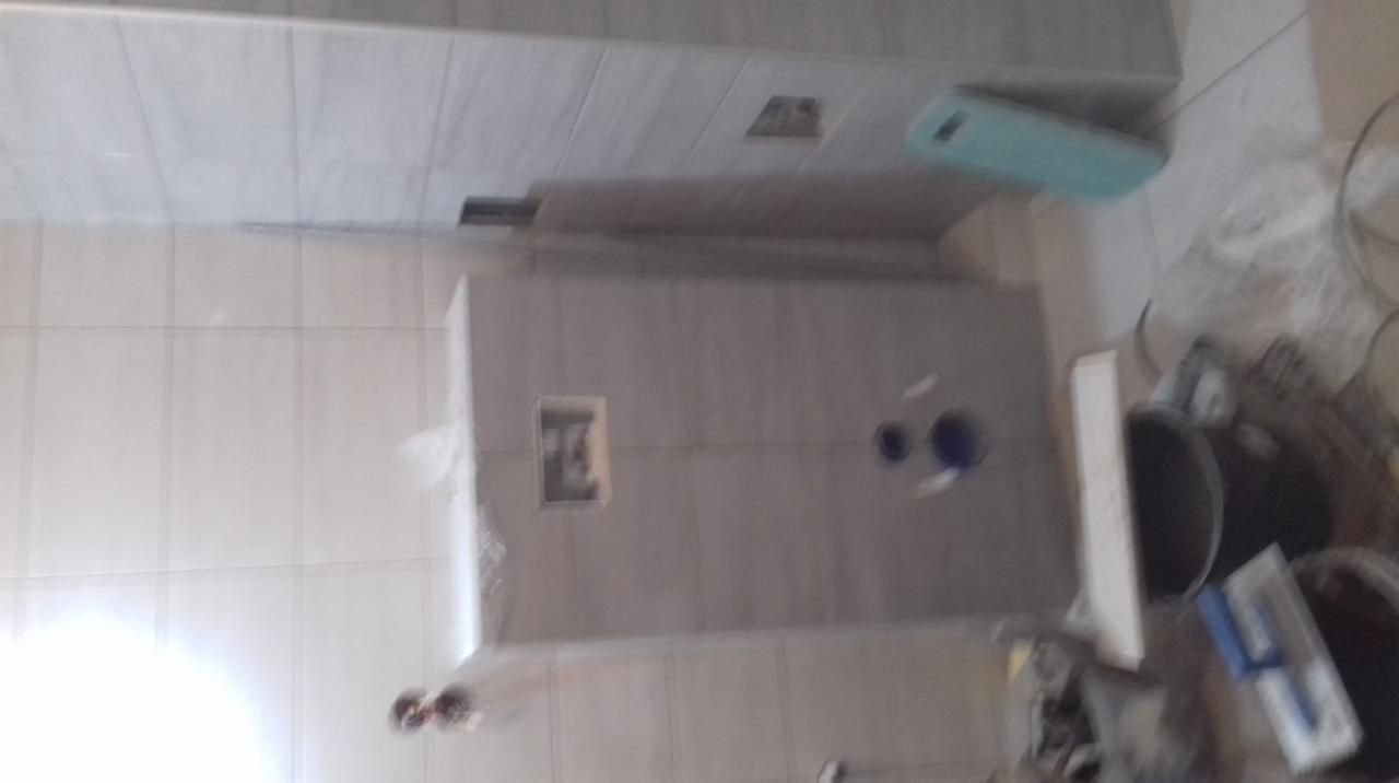 Niedokończona łazienka z szarymi płytkami, widoczne otwory na rury, narzędzia glazurnicze i wiadro z zaprawą w nieostrym ujęciu.