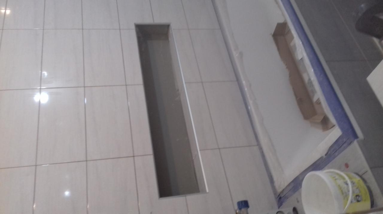 Ułożone jasne płytki podłogowe z wyciętym prostokątnym otworem, widoczny fragment brodzika z zabezpieczeniem folią i pojemnik z klejem w rogu.