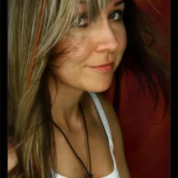 Portret uśmiechniętej kobiety z brązowymi oczami i blond włosami z pomarańczowymi pasemkami, ubrana w biały top na tle czerwonej ściany.