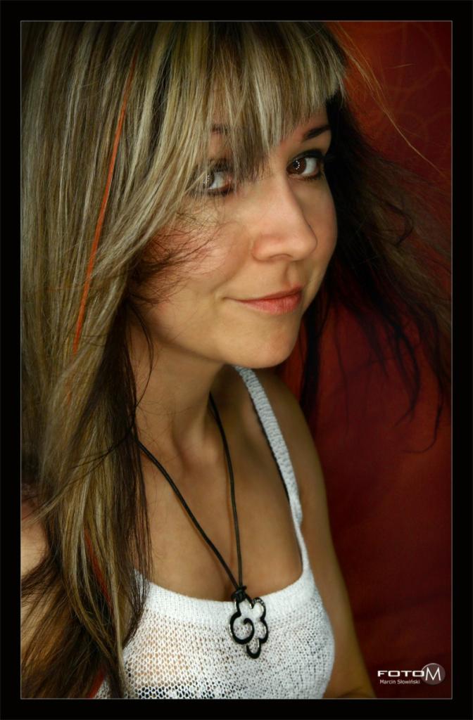 Portret uśmiechniętej kobiety z brązowymi oczami i blond włosami z pomarańczowymi pasemkami, ubrana w biały top na tle czerwonej ściany.