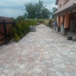 Ułożona kostka brukowa o nieregularnym kształcie w odcieniach szarości i beżu prowadząca do domu z kolumnami, widoczny fragment ogrodzenia, rząd tui i mężczyzna z pojemnikiem w tle