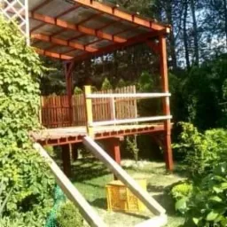 Drewniany taras na podwyższeniu z zadaszeniem, otoczony zielenią, z widocznymi schodami i skrzynką narzędziową.