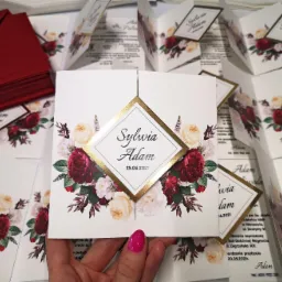 Stos eleganckich, otwartych zaproszeń ślubnych z motywem kwiatowym i złotym akcentem, ułożonych na białym tle, z widocznymi czerwonymi kopertami i kobiecą dłonią z różowym manicure.