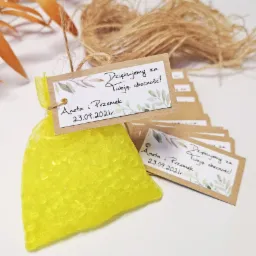 Żółta sakiewka z drobnymi koralikami i dołączona kartka z podziękowaniem za obecność, obok leżą dodatkowe kartki z podziękowaniami, całość na białym tle z dekoracyjnymi gałązkami.