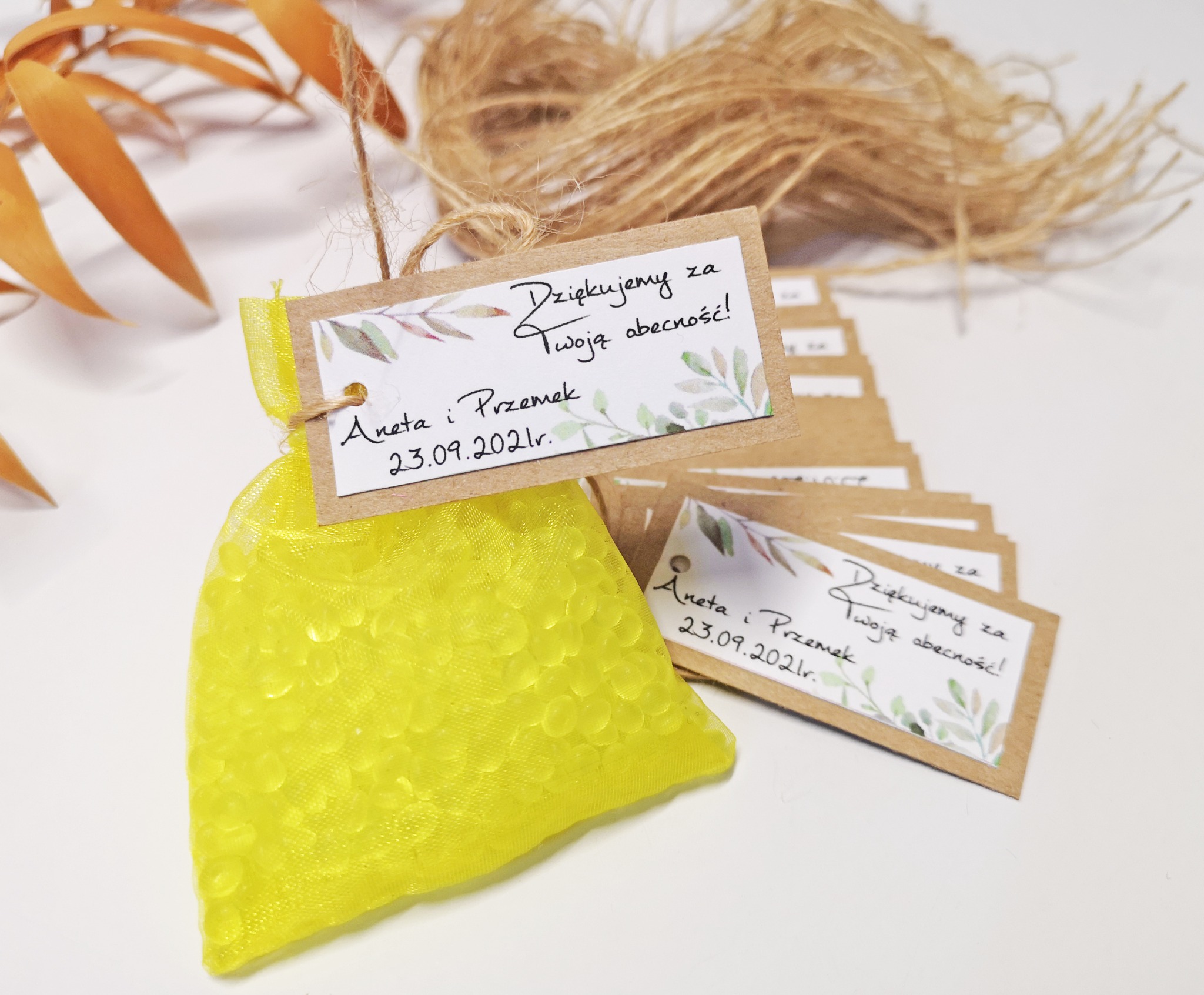 Żółta sakiewka z drobnymi koralikami i dołączona kartka z podziękowaniem za obecność, obok leżą dodatkowe kartki z podziękowaniami, całość na białym tle z dekoracyjnymi gałązkami.