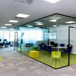 Nowoczesne biuro z przeszkloną salą konferencyjną, widoczne biurka i krzesła w tle, szaro-zielona wykładzina podłogowa.