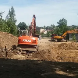 Pomarańczowa koparka gąsienicowa marki Atlas na placu budowy, w tle żółta koparka Hyundai i budynek gospodarczy.