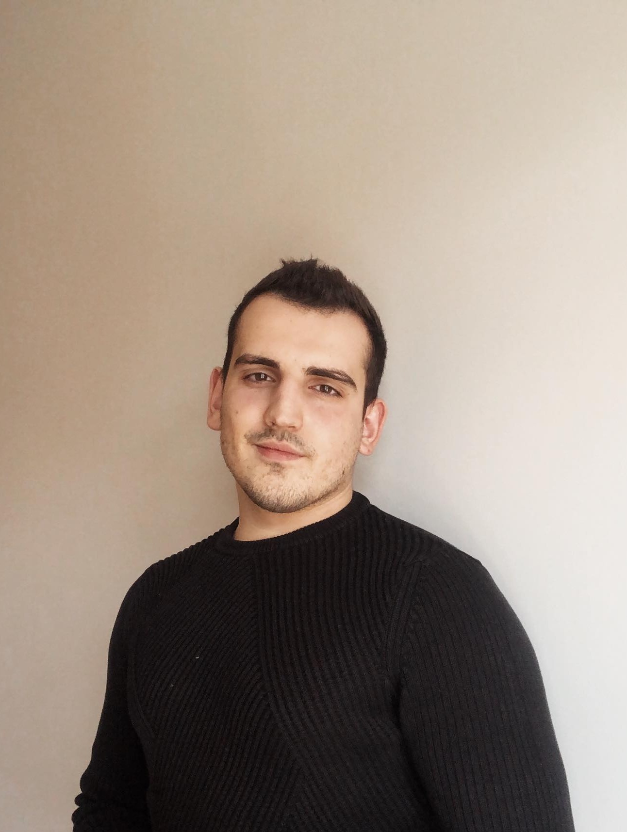 Młody mężczyzna w czarnym swetrze, uśmiechający się lekko na jasnym tle, portret biznesowy.