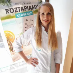 Uśmiechnięta blondynka w białym uniformie na tle plakatu z napisem 'Roztapiamy kilogramy' i skalą wagi.