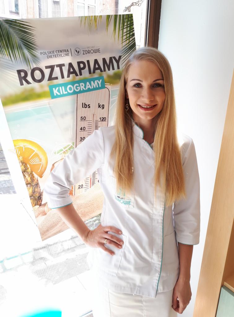 Uśmiechnięta blondynka w białym uniformie na tle plakatu z napisem 'Roztapiamy kilogramy' i skalą wagi.