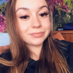 Portret uśmiechniętej brunetki z długimi sztucznymi rzęsami na tle ściany z motywem kwiatowym.