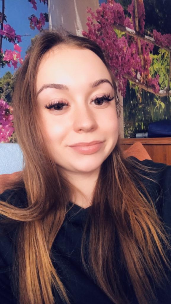 Portret uśmiechniętej brunetki z długimi sztucznymi rzęsami na tle ściany z motywem kwiatowym.