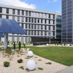 Zewnętrzne panele słoneczne zamontowane na fasadzie budynku biurowego i na postumentach w ogrodzie, oświetlające teren za pomocą kulistych lamp.