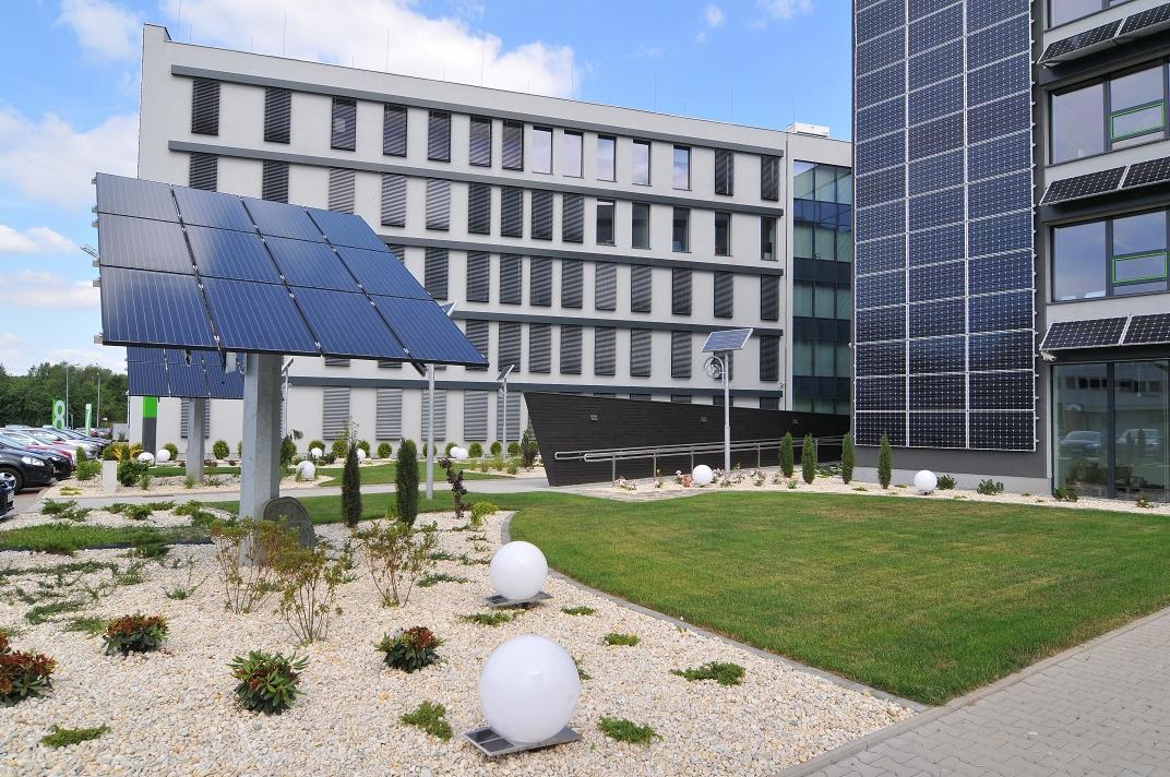 Zewnętrzne panele słoneczne zamontowane na fasadzie budynku biurowego i na postumentach w ogrodzie, oświetlające teren za pomocą kulistych lamp.