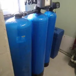 Trzy pionowe, niebieskie zbiorniki systemu uzdatniania wody z elektronicznymi panelami sterującymi, ustawione w pomieszczeniu z betonową podłogą i zieloną ścianą.