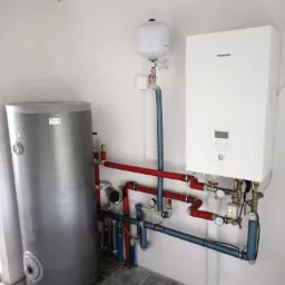 Nowoczesna kotłownia z białym piecem gazowym Panasonic, szarym zasobnikiem ciepłej wody i systemem rur w kolorach czerwonym i niebieskim, zamontowana w surowym, białym pomieszczeniu.