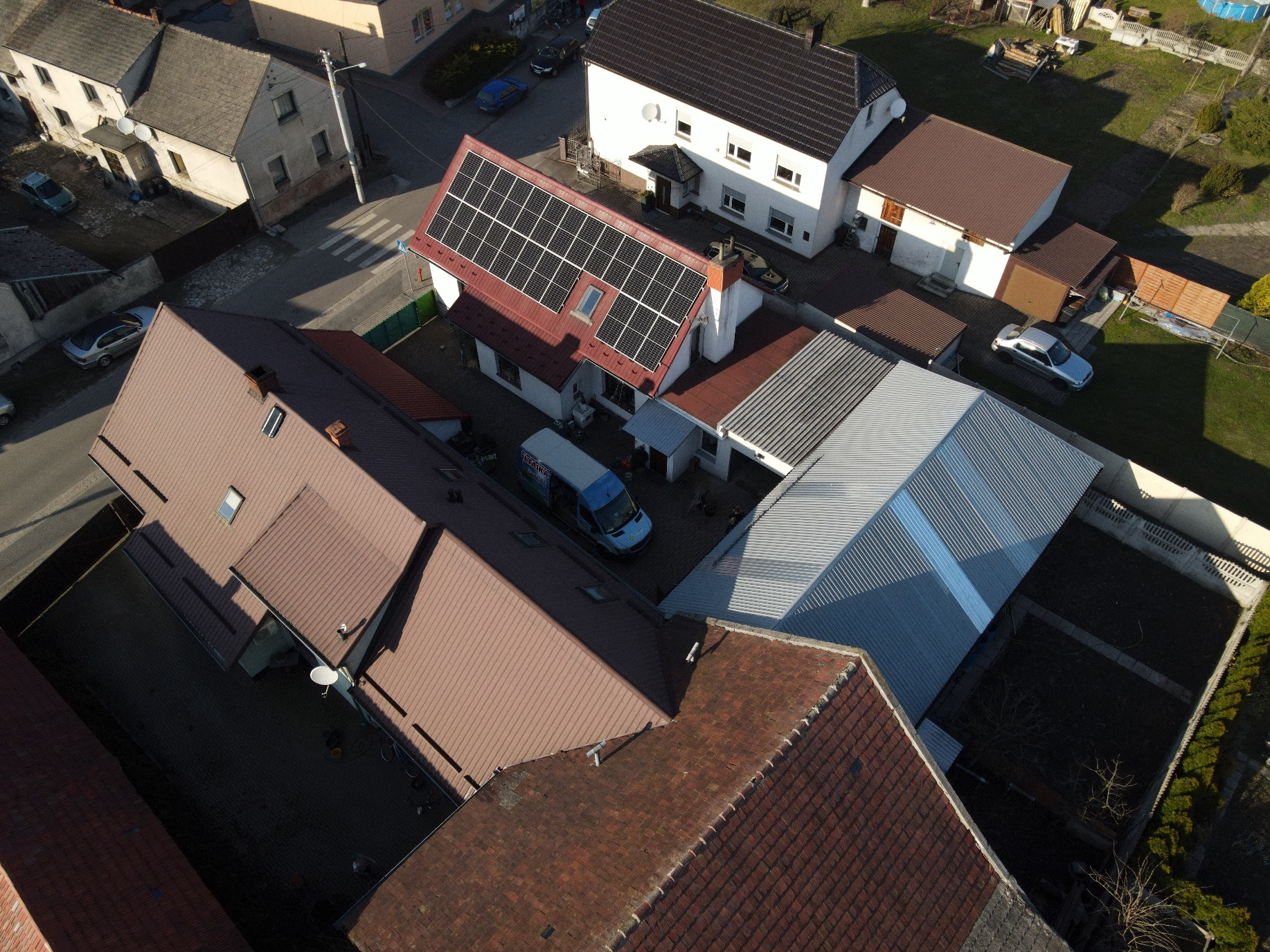 Widok z lotu ptaka na dom z panelami słonecznymi na czerwonym dachu, obok zaparkowany bus firmy instalującej panele.