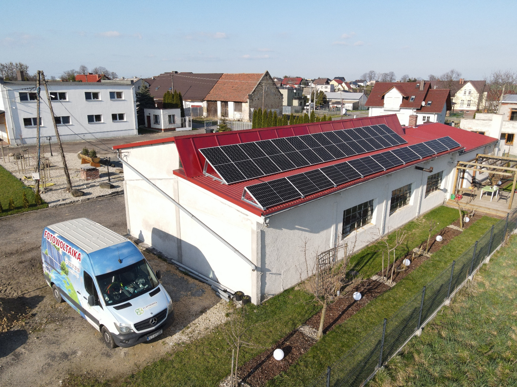 Instalacja paneli słonecznych na czerwonym dachu budynku w Tychach, widok z góry. Na pierwszym planie firmowy bus z reklamą fotowoltaiki.