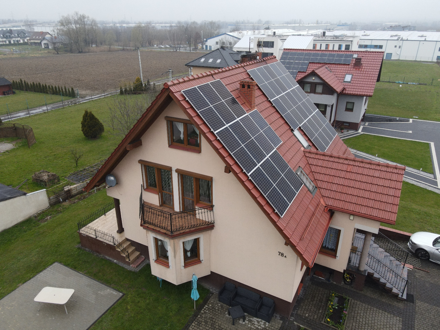 Widok z góry na dom jednorodzinny z zamontowanymi panelami słonecznymi na czerwonym dachu, widoczne również inne domy i zabudowania w tle.