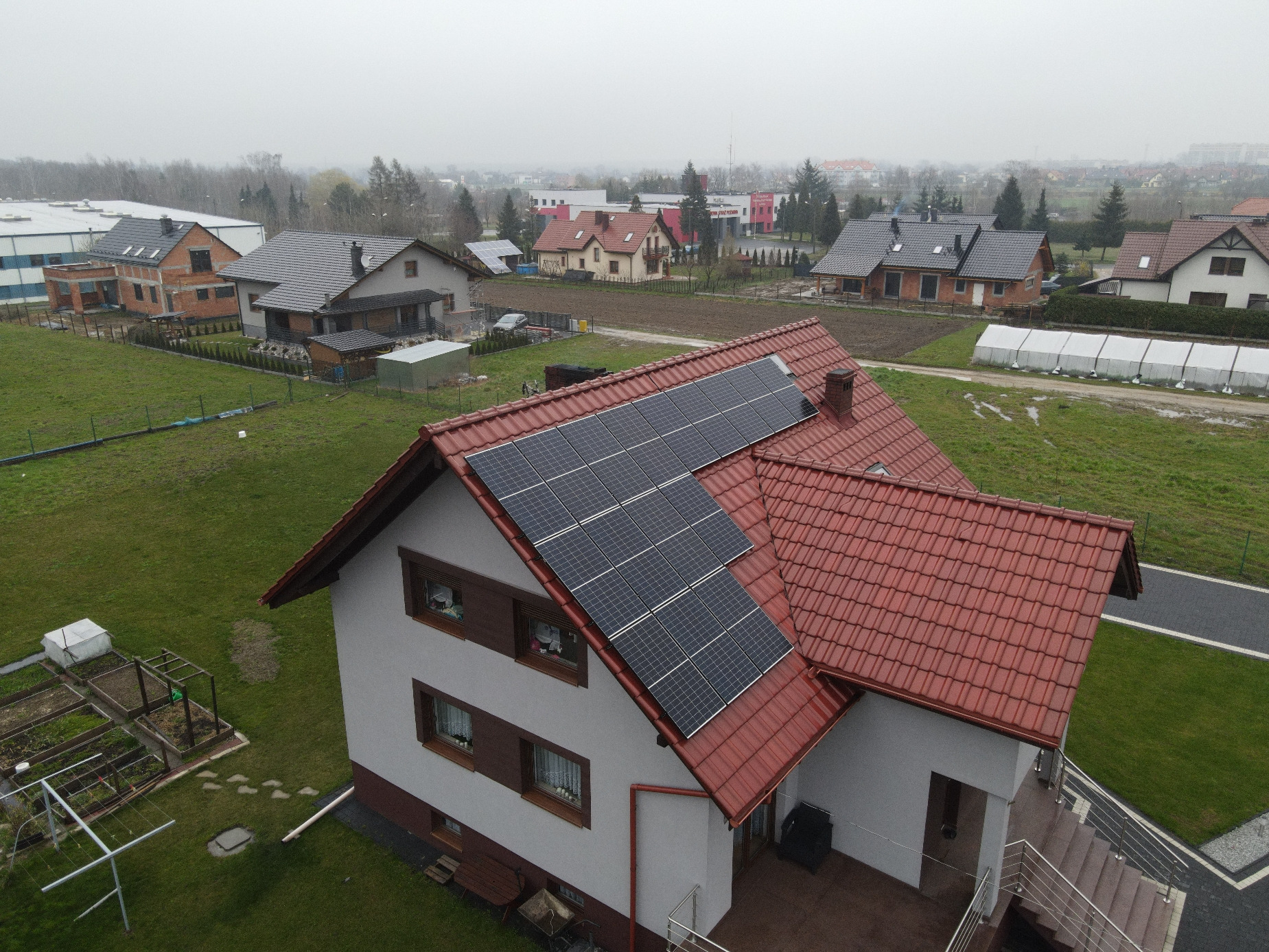 Widok z góry na dom jednorodzinny z czerwoną dachówką i zamontowanymi panelami słonecznymi; w tle zabudowa mieszkalna i pola uprawne, pochmurne niebo.