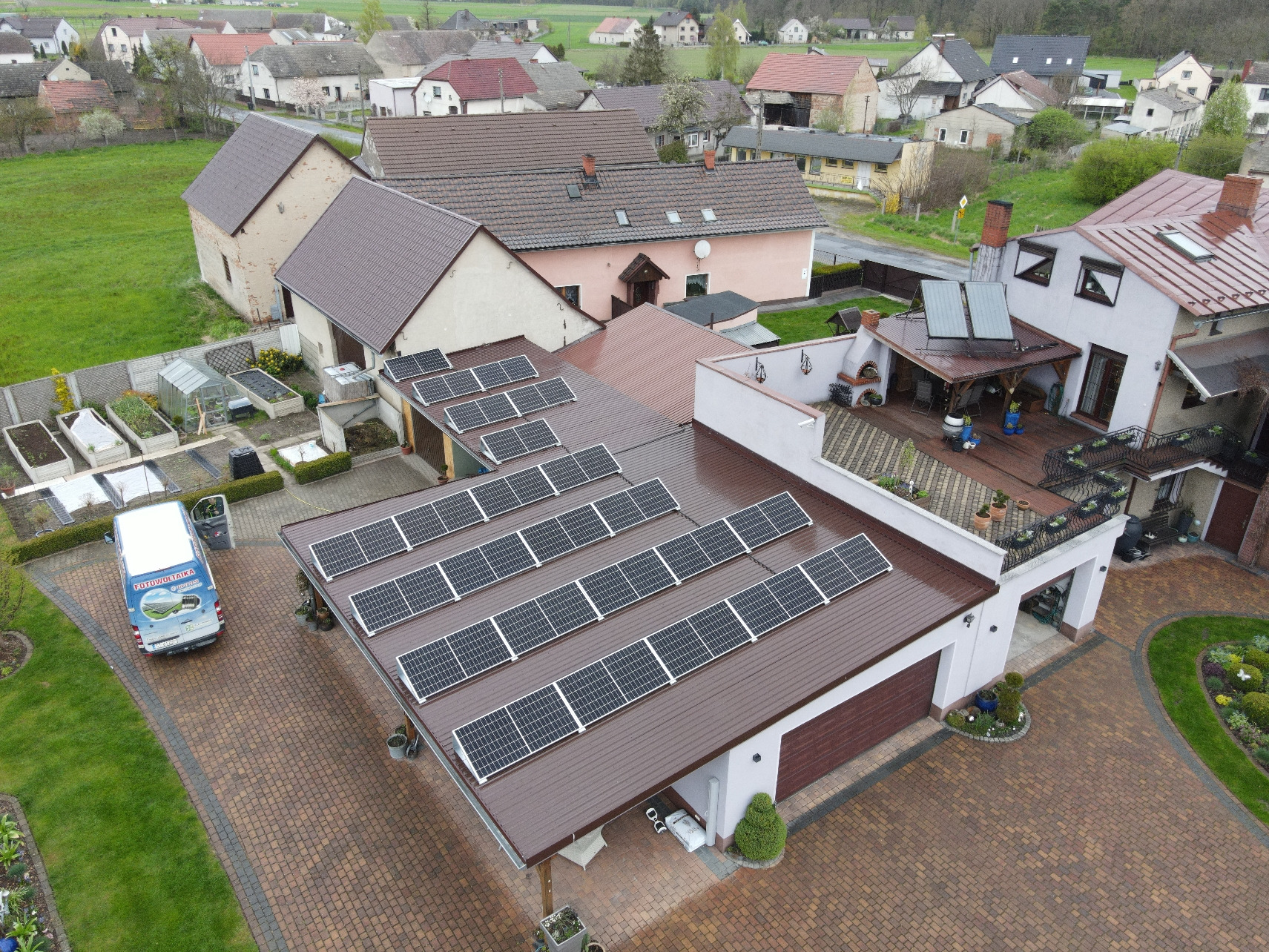 Widok z góry na dom z brązowym dachem pokrytym panelami słonecznymi; na podjeździe stoi bus z logo firmy instalującej systemy fotowoltaiczne.