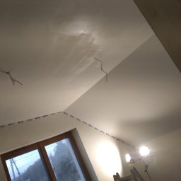 FACH-BUD Rafał Bobek - Sufit po szpachlowaniu, z widocznymi śladami po taśmie zabezpieczającej krawędzie, oświetlony lampami budowlanymi, okno w tle.