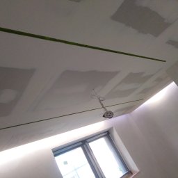 FACH-BUD Rafał Bobek - Sufit w trakcie prac wykończeniowych: widoczne płyty kartonowo-gipsowe, miejsca szpachlowane, zielona taśma malarska wyznaczająca linie oraz wisząca oprawa oświetleniowa. W tle okno.