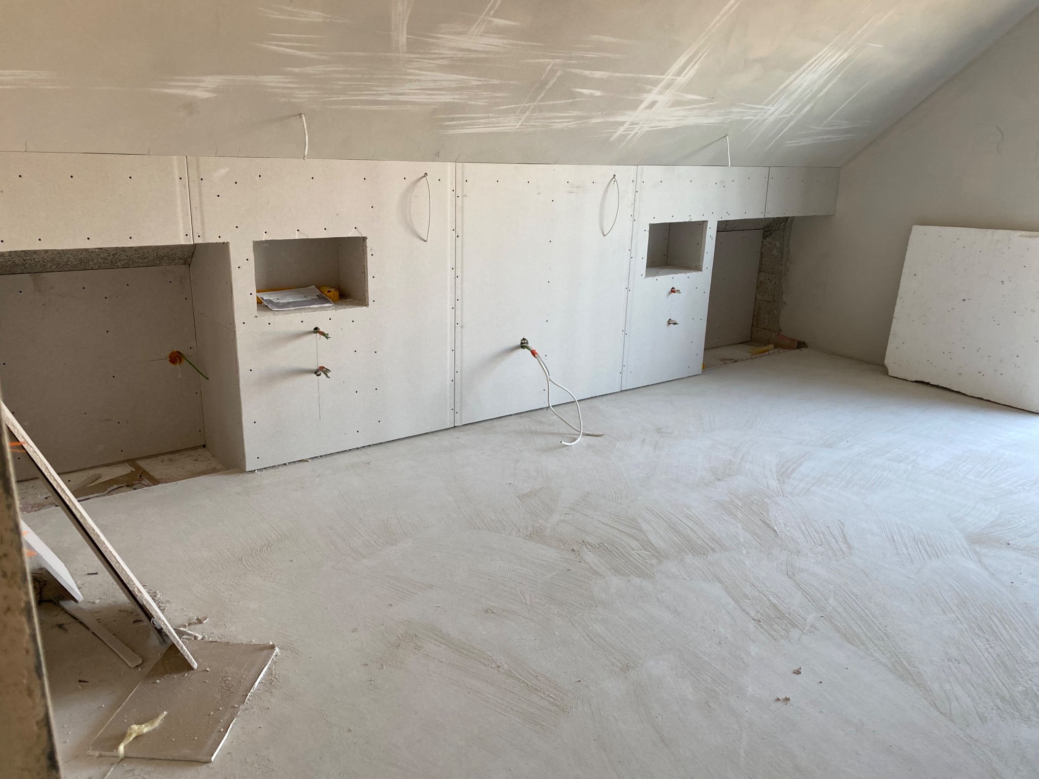 Poddasze w trakcie wykańczania, ściany i sufit pokryte płytami kartonowo-gipsowymi, widoczne otwory pod instalacje elektryczne i wnęki, surowa wylewka na podłodze.