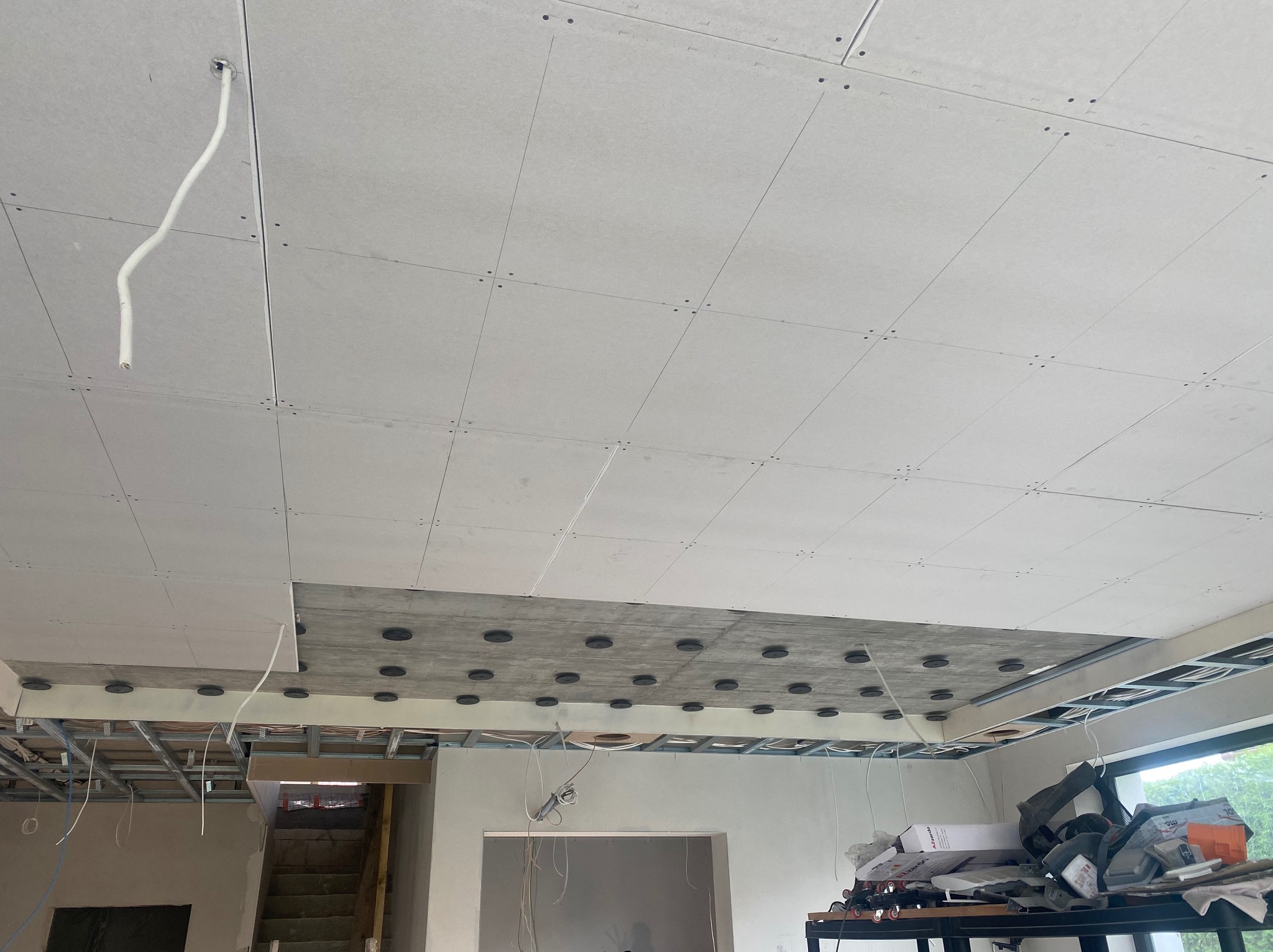 Montaż sufitu podwieszanego z płyt kartonowo-gipsowych, widoczne elementy konstrukcji nośnej i otwory wentylacyjne w betonie, przewody elektryczne wystające z sufitu.