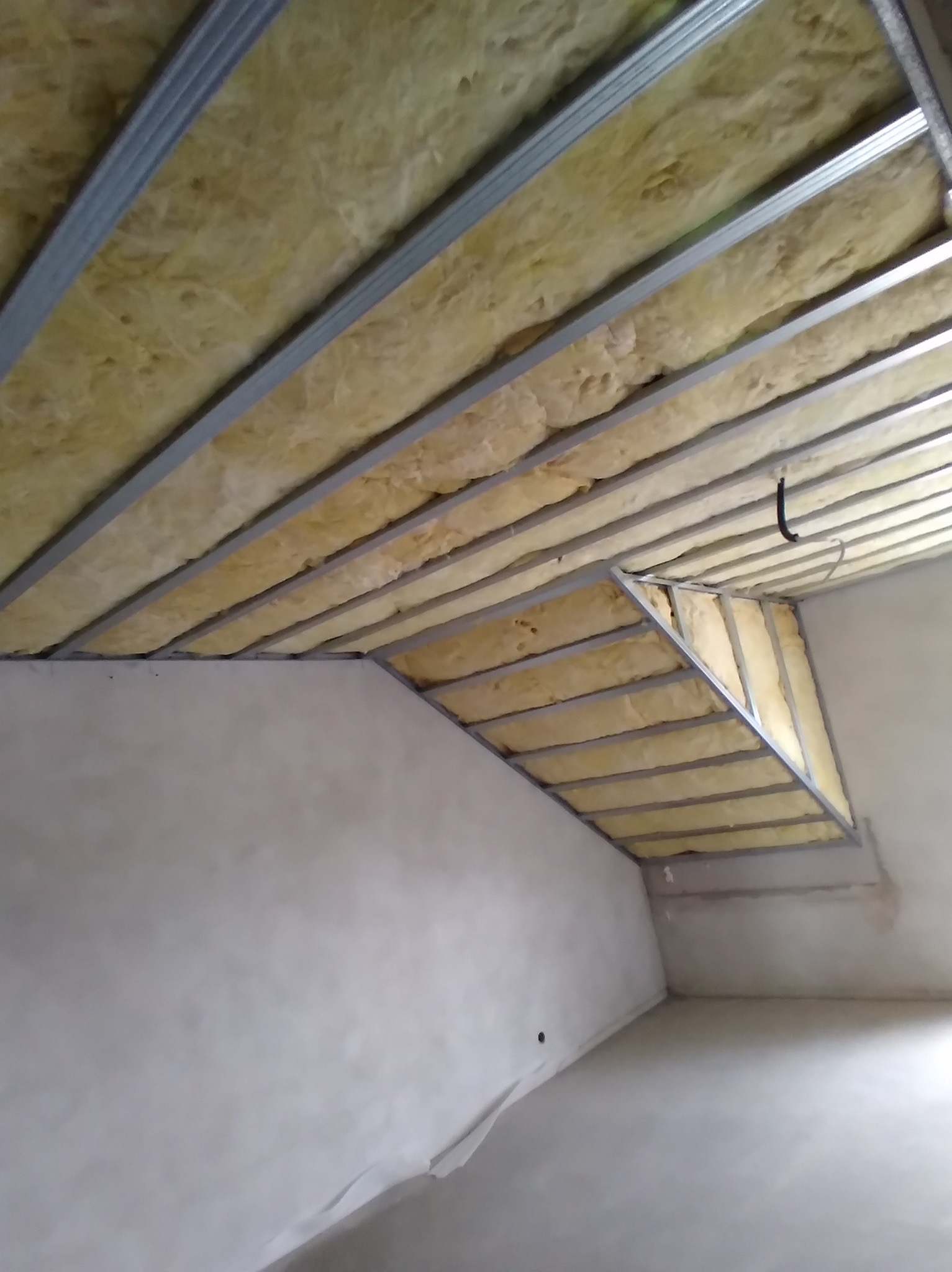 Montaż izolacji z wełny mineralnej w metalowym stelażu sufitu podwieszanego na poddaszu, widoczne nieotynkowane ściany.