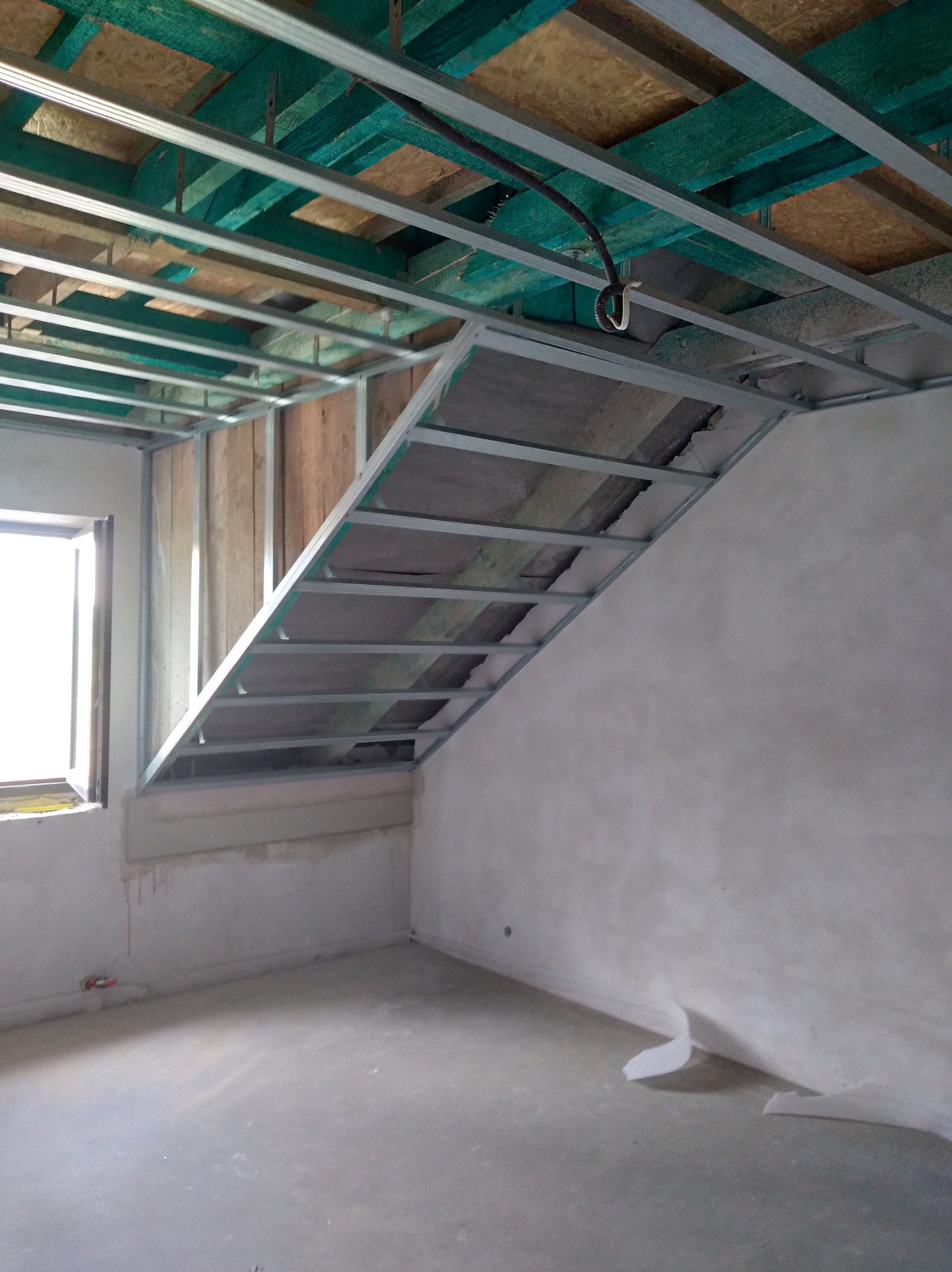 Wnętrze w trakcie prac wykończeniowych: metalowa konstrukcja sufitu podwieszanego z widoczną instalacją elektryczną, fragment ściany z surowym tynkiem, okno z widocznym parapetem, surowa podłoga.