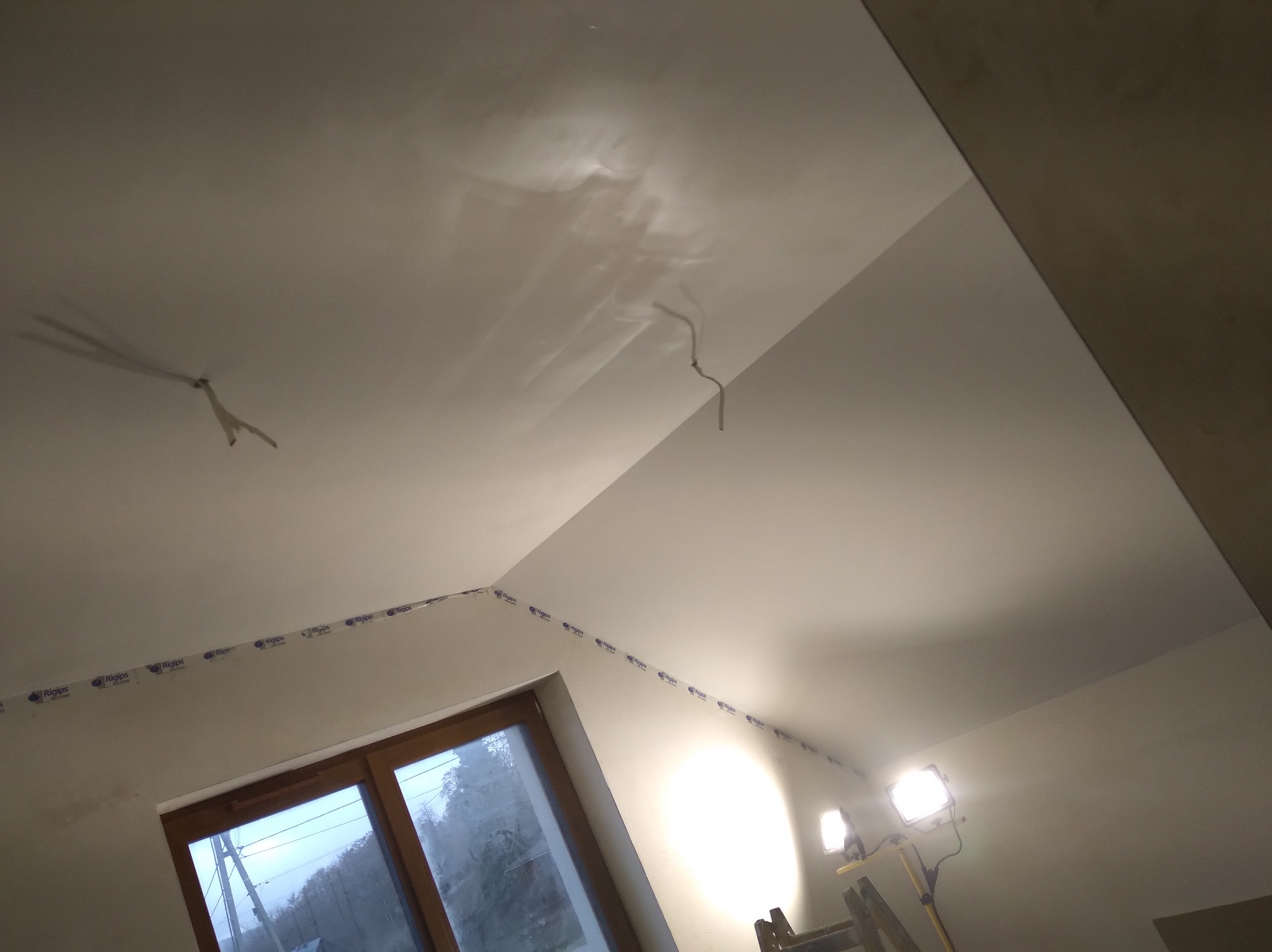 Sufit po szpachlowaniu, z widocznymi śladami po taśmie zabezpieczającej krawędzie, oświetlony lampami budowlanymi, okno w tle.