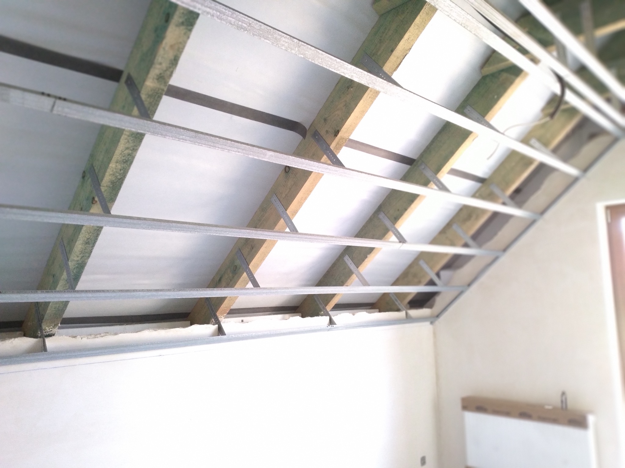 Szkielet sufitu podwieszanego z profili metalowych i drewnianych belek, wypełniony białą wełną mineralną, widok z dołu na poddaszu.