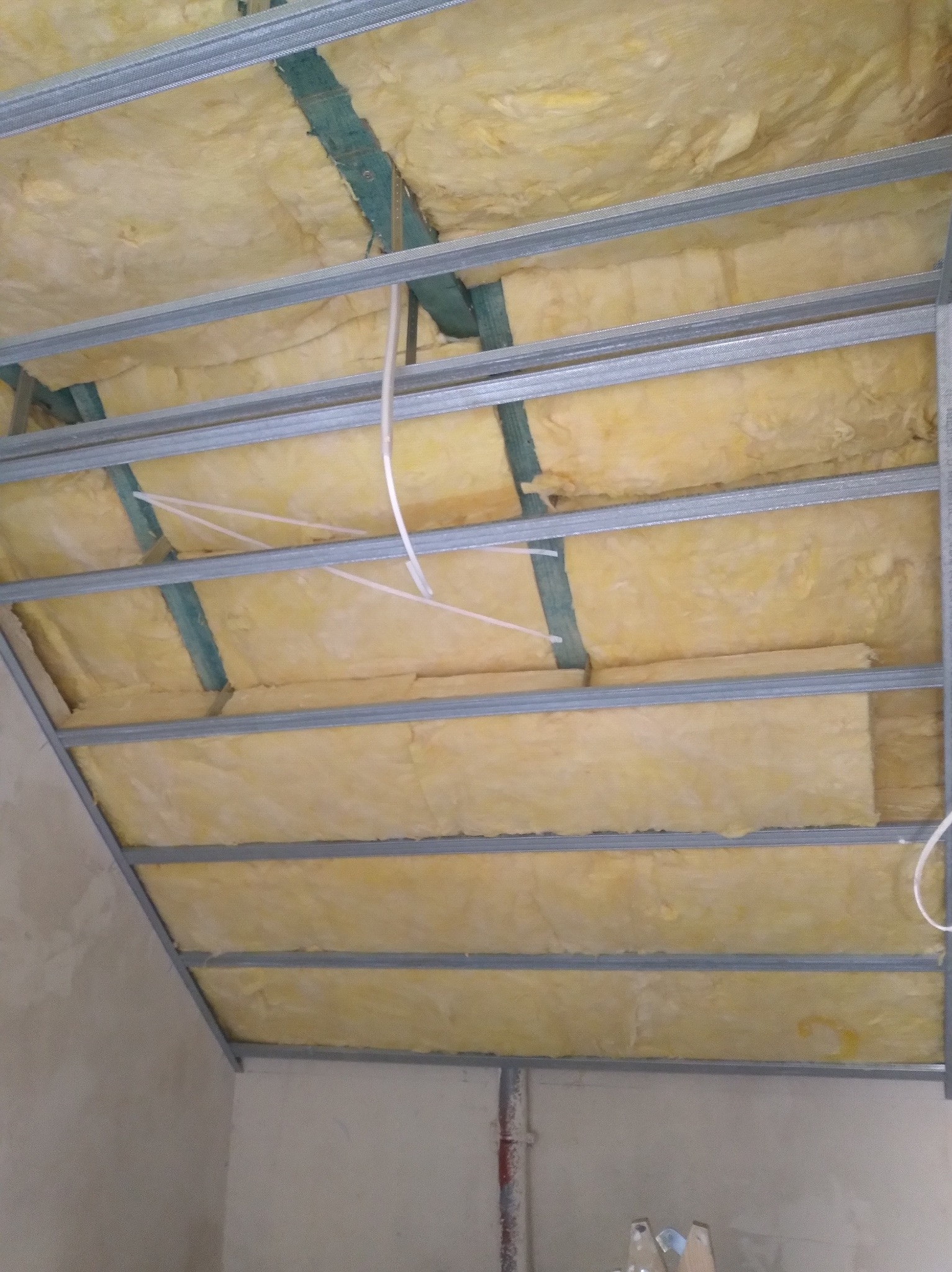 Montaż izolacji z wełny mineralnej w metalowym stelażu sufitu podwieszanego, widok od dołu, widoczne elementy instalacji elektrycznej.