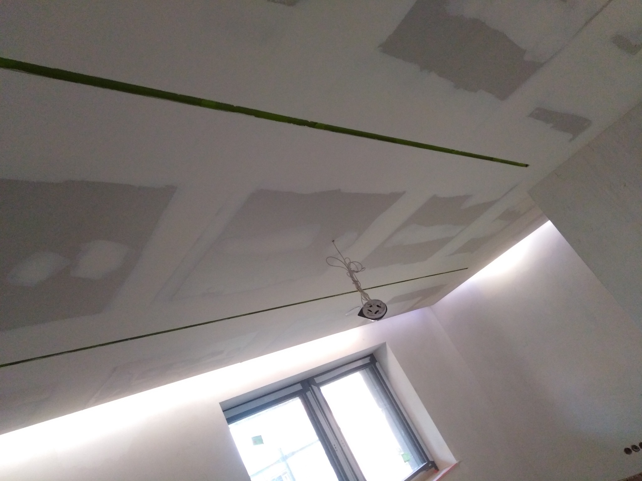 Sufit w trakcie prac wykończeniowych: widoczne płyty kartonowo-gipsowe, miejsca szpachlowane, zielona taśma malarska wyznaczająca linie oraz wisząca oprawa oświetleniowa. W tle okno.