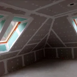 Poddasze w trakcie wykańczania, widoczne płyty na ścianach i suficie, spoiny wypełnione masą, okna dachowe z widokiem na las.