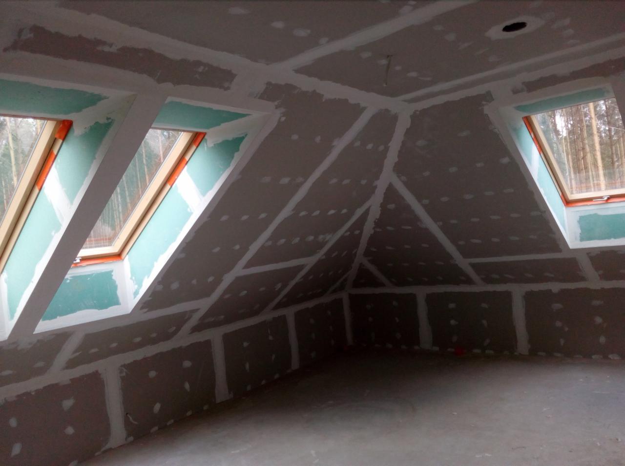 Poddasze w trakcie wykańczania, widoczne płyty na ścianach i suficie, spoiny wypełnione masą, okna dachowe z widokiem na las.
