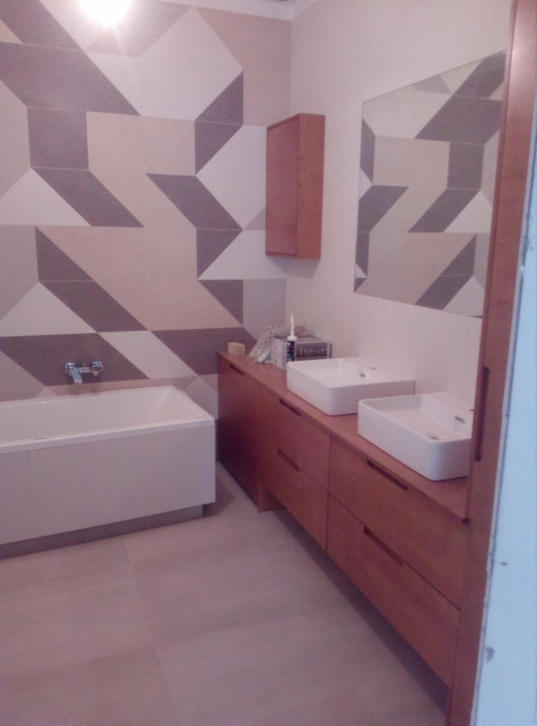 Wykończona łazienka z geometrycznym wzorem płytek na ścianie, dwiema białymi umywalkami nablatowymi na drewnianej szafce z szufladami i wanną.