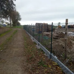 Długa linia ogrodzenia z siatki, wsparta na betonowej podmurówce, biegnąca wzdłuż polnej drogi, z zaparkowanymi skuterami i stertą drewna w tle.