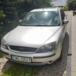 Srebrny Ford Focus zaparkowany na kostce brukowej przed zielonymi krzewami w słoneczny dzień, widoczne polskie tablice rejestracyjne CWL ER21.